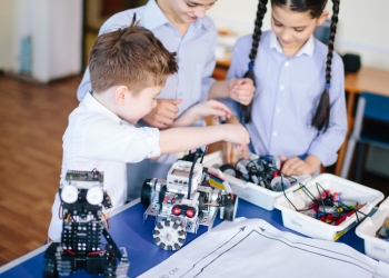 Excited little kid and two smiling teen friends construct robotic toys while playing during their leisure time in technical workshop. Intelligent activity.